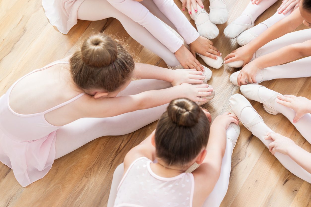 Of a group of ballerinas, we can see only their hands and feet, a few legs and a couple of backs and heads as they sit on the floor in a circle with their legs extended and their pointed toes together while each touching their toes.
